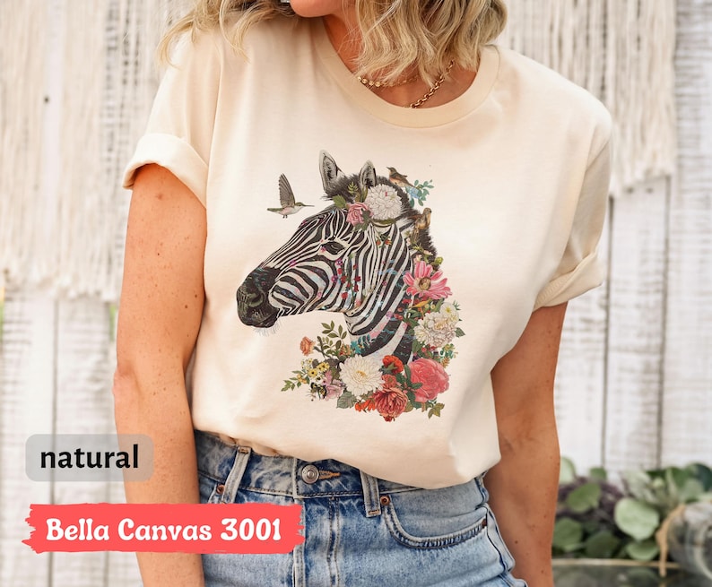 May include: A natural colored t-shirt with a zebra print design. The zebra is surrounded by flowers and birds. The text "Bella Canvas 3001" is printed on the bottom of the shirt.