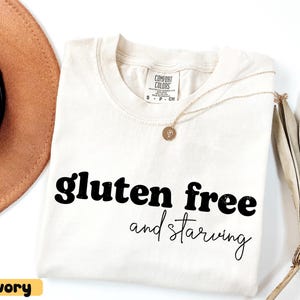 Puede incluir: Camiseta de color marfil con las palabras "gluten free and starving" en letras negras. La camiseta tiene cuello redondo y mangas cortas. Un collar dorado con un colgante est&aacute; sobre la camiseta. Un sombrero marr&oacute;n es visible en el fondo.