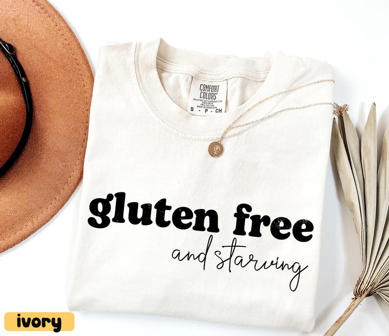 Puede incluir: Camiseta de color marfil con las palabras "gluten free and starving" en letras negras. La camiseta tiene cuello redondo y mangas cortas. Un collar dorado con un colgante est&aacute; sobre la camiseta. Un sombrero marr&oacute;n es visible en el fondo.