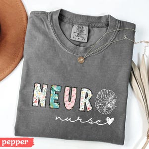 Puede incluir: Una camiseta gris con las palabras "NEURO nurse" en un dise&ntilde;o floral. La palabra "NEURO" est&aacute; en letras coloridas, y un gr&aacute;fico de cerebro est&aacute; a la derecha. Un collar plateado con un colgante est&aacute; sobre la camiseta.