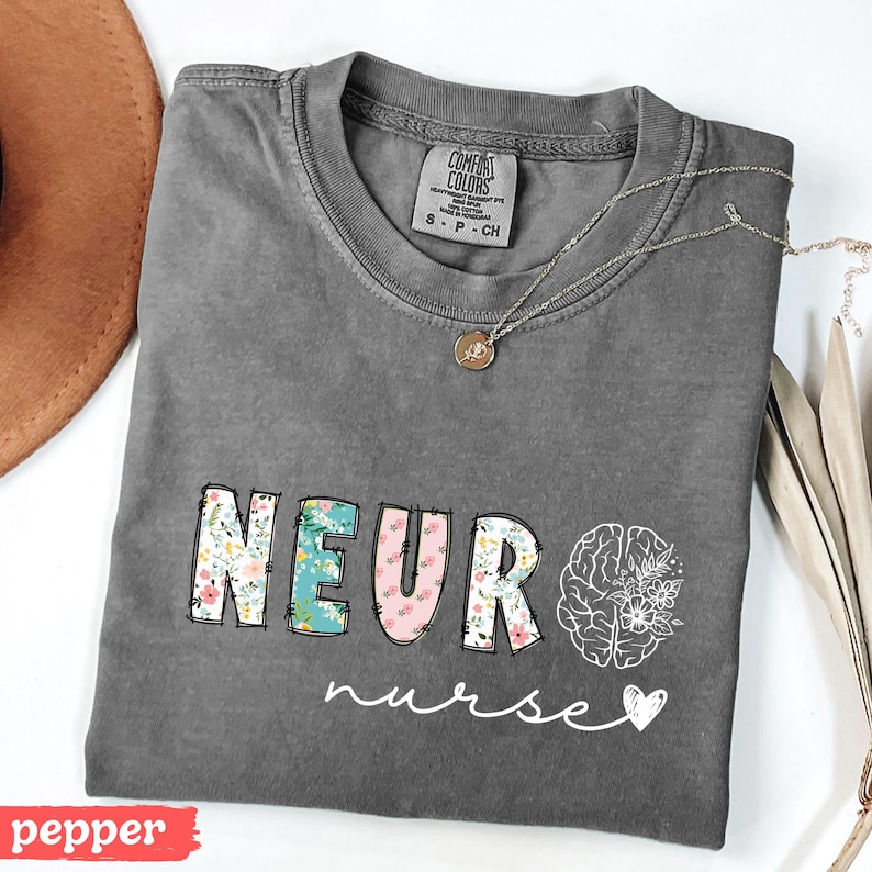 Puede incluir: Una camiseta gris con las palabras "NEURO nurse" en un dise&ntilde;o floral. La palabra "NEURO" est&aacute; en letras coloridas, y un gr&aacute;fico de cerebro est&aacute; a la derecha. Un collar plateado con un colgante est&aacute; sobre la camiseta.