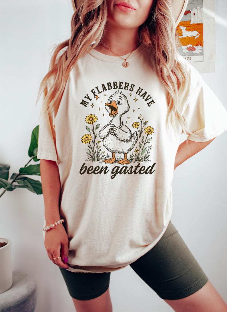 May include: Cream-colored t-shirt featuring a cartoon duck with the text "MY FLABBERS HAVE been gasted". The duck is surrounded by flowers and stars. The shirt is a casual, comfortable style.