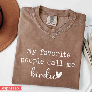 May include: Espresso-coloured t-shirt featuring the text "my favourite people call me birdie" in white script. A silver necklace with a small pendant lies on the shirt. The shirt has a crew neck and a Comfort Colors label.