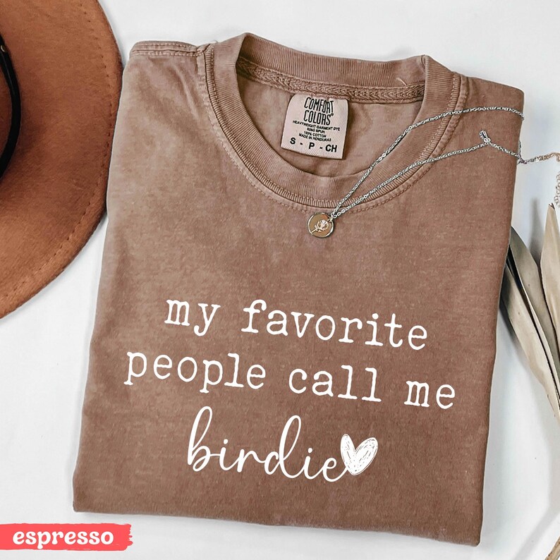 May include: Espresso-coloured t-shirt featuring the text "my favourite people call me birdie" in white script. A silver necklace with a small pendant lies on the shirt. The shirt has a crew neck and a Comfort Colors label.