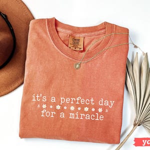 May include: A coral-colored t-shirt with the text "it's a perfect day for a miracle" in white lettering. The shirt has a distressed look and is decorated with small white flower designs.