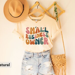 May include: A beige t-shirt with the text "SMALL BUSINESS OWNER" in colorful, retro-style lettering. The shirt is hanging on a wooden hanger and has a natural color.