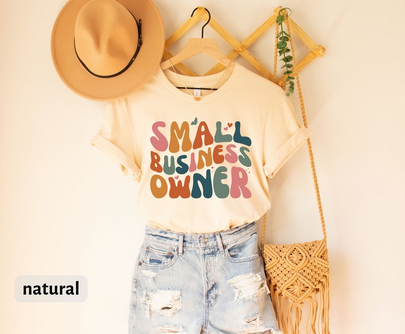 May include: A beige t-shirt with the text "SMALL BUSINESS OWNER" in colorful, retro-style lettering. The shirt is hanging on a wooden hanger and has a natural color.