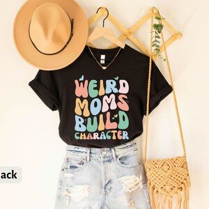 May include: Black t-shirt with the text "Weird Moms Build Character" in colourful letters. The shirt is styled with a straw hat, denim shorts, and a macrame bag.