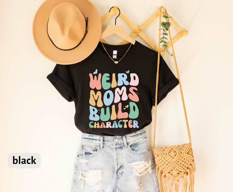 May include: Black t-shirt with the text "Weird Moms Build Character" in colourful letters. The shirt is styled with a straw hat, denim shorts, and a macrame bag.