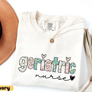 Pode incluir: Camiseta cor marfim com as palavras "Geriatric nurse" em um design floral e de cora&ccedil;&atilde;o. A camiseta tem gola redonda e mangas curtas. Um colar de ouro com um pingente est&aacute; drapeado na camiseta. Um chap&eacute;u marrom &eacute; vis&iacute;vel no fundo.