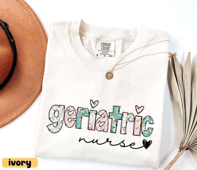 Pode incluir: Camiseta cor marfim com as palavras "Geriatric nurse" em um design floral e de cora&ccedil;&atilde;o. A camiseta tem gola redonda e mangas curtas. Um colar de ouro com um pingente est&aacute; drapeado na camiseta. Um chap&eacute;u marrom &eacute; vis&iacute;vel no fundo.