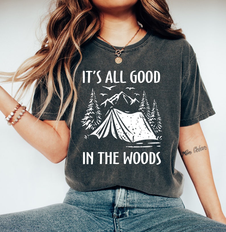 May include: A dark gray t-shirt with a white graphic of a tent in the woods. The text "It's all good in the woods" is printed on the shirt.