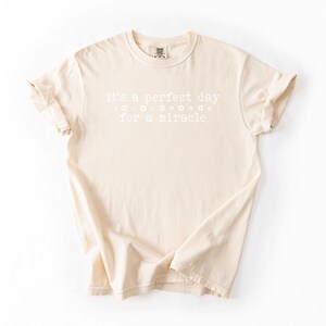 May include: A beige t-shirt with the text "It's a perfect day for a miracle" printed in white.