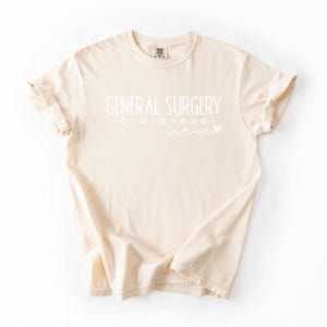 Może przedstawiać: Beżowa koszulka z napisem "GENERAL SURGERY" w białym kolorze. Tekst jest otoczony małymi białymi kwiatami i sercem.