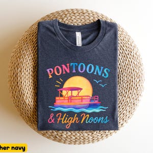 May include: Heather navy t-shirt with a colorful graphic. The design features a pontoon boat on water, a large yellow sun, and the text "Pontoons & High Noons" in a rainbow gradient. The shirt is displayed on a woven surface.