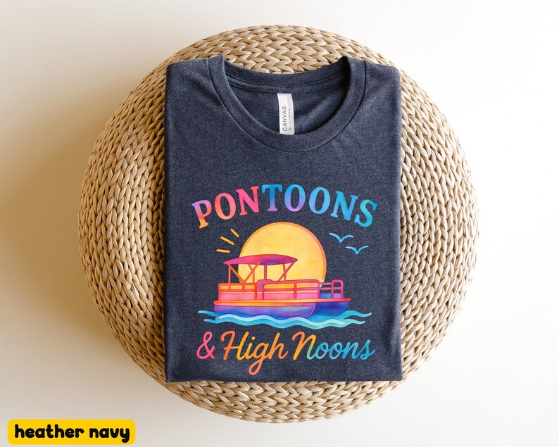 May include: Heather navy t-shirt with a colorful graphic. The design features a pontoon boat on water, a large yellow sun, and the text "Pontoons & High Noons" in a rainbow gradient. The shirt is displayed on a woven surface.
