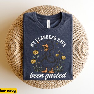 May include: Heather navy t-shirt with a graphic of a duck with the text "MY FLABBERS HAVE been gasted". The duck is surrounded by yellow flowers and stars. The shirt is displayed on a woven surface.