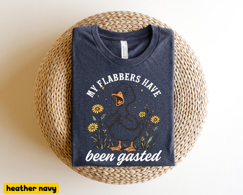 May include: Heather navy t-shirt with a graphic of a duck with the text "MY FLABBERS HAVE been gasted". The duck is surrounded by yellow flowers and stars. The shirt is displayed on a woven surface.