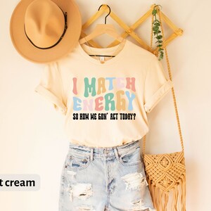 May include: A soft cream colored t-shirt with the text "I Match Energy So How We Gon' Act Today?" in colorful, wavy letters.