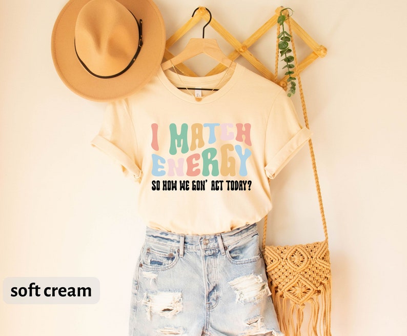 May include: A soft cream colored t-shirt with the text "I Match Energy So How We Gon' Act Today?" in colorful, wavy letters.