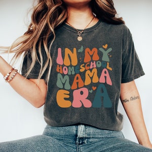 May include: A dark gray t-shirt with the text "In My Homeschool Mama Era" in colorful, retro-style lettering. The text is surrounded by small hearts and stars.