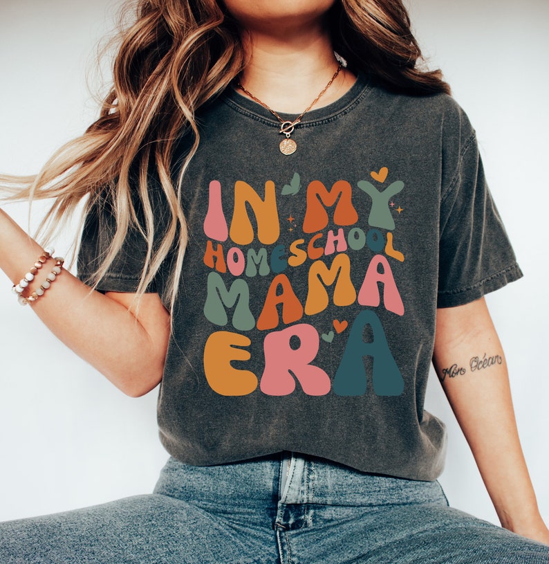 May include: A dark gray t-shirt with the text "In My Homeschool Mama Era" in colorful, retro-style lettering. The text is surrounded by small hearts and stars.
