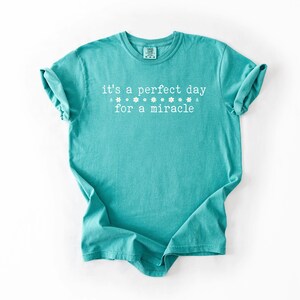 May include: Teal blue t-shirt with white text that reads "it's a perfect day for a miracle" with a row of white stars above the text.
