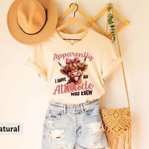 May include: A cream colored t-shirt with a cartoon Highland cow with a pink bow. The text on the shirt reads "Apparently I have an attitude. Who knew."