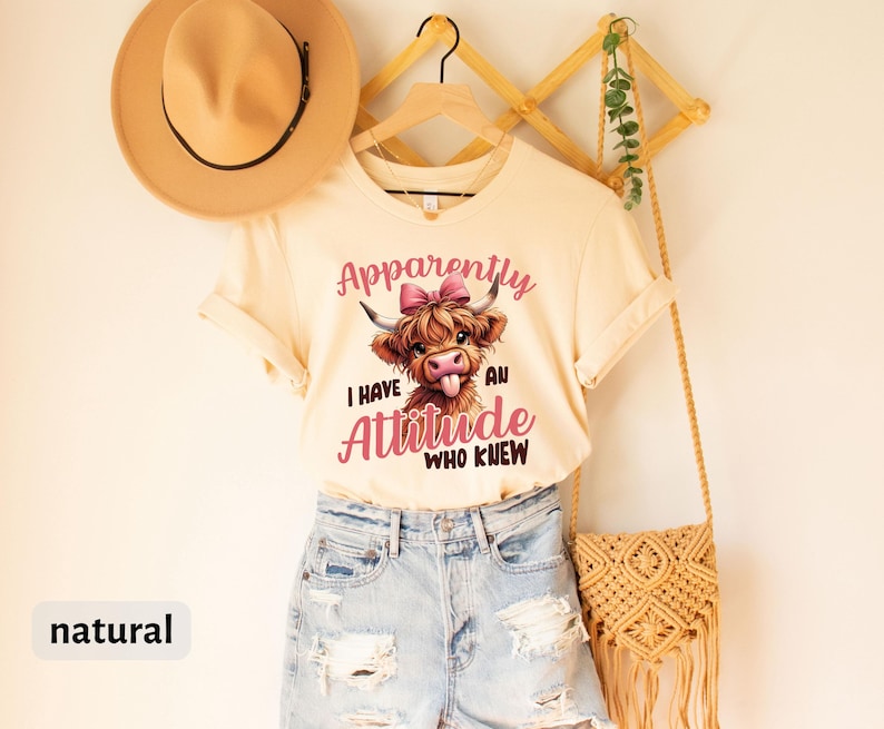 May include: A cream colored t-shirt with a cartoon Highland cow with a pink bow. The text on the shirt reads "Apparently I have an attitude. Who knew."