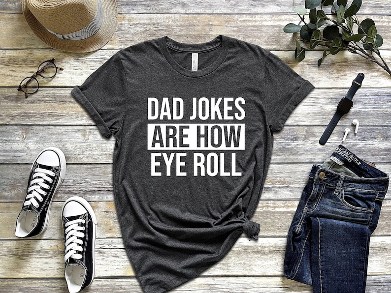 May include: A dark gray t-shirt with white text that reads "DAD JOKES ARE HOW EYE ROLL". The shirt is laid flat on a wooden surface with a pair of black sneakers, a straw hat, a pair of glasses, a watch, and a pair of earbuds.