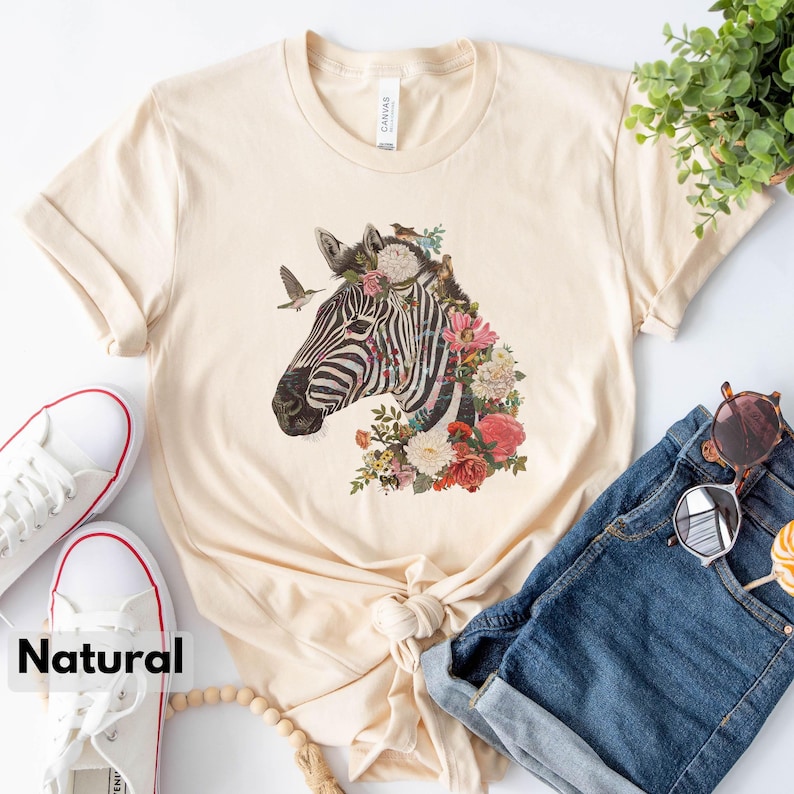 May include: A beige t-shirt with a graphic of a zebra's head with flowers and birds. The zebra is black and white and the flowers are pink, red, and yellow.