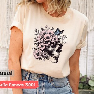 May include: A beige t-shirt with a black and white skull graphic with pink flowers and a blue butterfly on top of the skull. The text "Bella Canvas 3001" and "natural" are visible on the bottom of the image.