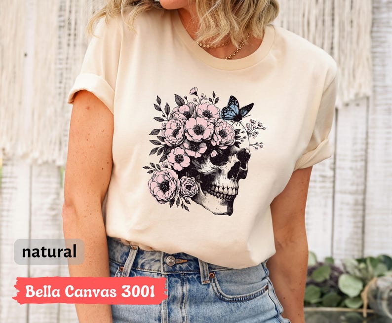 May include: A beige t-shirt with a black and white skull graphic with pink flowers and a blue butterfly on top of the skull. The text "Bella Canvas 3001" and "natural" are visible on the bottom of the image.