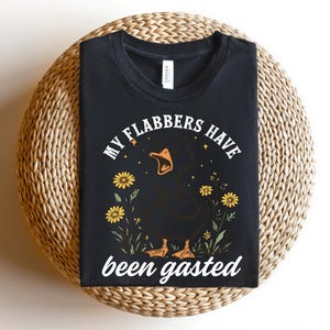 May include: Black t-shirt with a graphic of a black duck with an open beak, surrounded by yellow flowers and the text "MY FLABBERS HAVE been gasted". The shirt is displayed on a woven, circular surface.