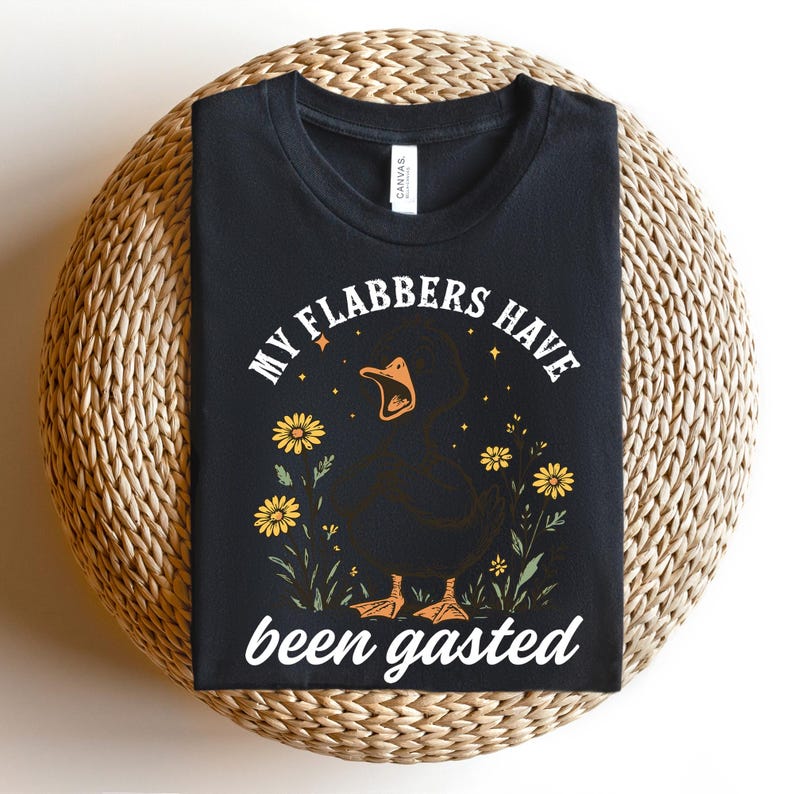 May include: Black t-shirt with a graphic of a black duck with an open beak, surrounded by yellow flowers and the text "MY FLABBERS HAVE been gasted". The shirt is displayed on a woven, circular surface.