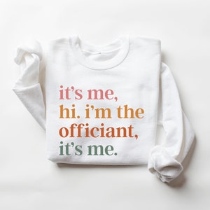 May include: White sweatshirt with the text "it's me, hi. i'm the officiant, it's me." printed in colourful letters.