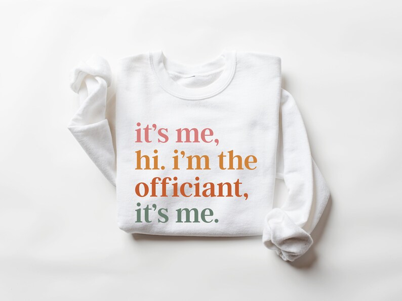May include: White sweatshirt with the text "it's me, hi. i'm the officiant, it's me." printed in colourful letters.