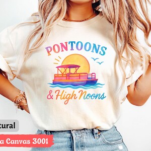 May include: Cream-colored t-shirt with a colorful graphic of a pontoon boat on water, a sun, and the words "Pontoons & High Noons." The shirt is made of Bella Canvas 3001 material.
