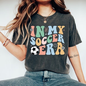Może przedstawiać: Szary t-shirt z napisem "IN MY SOCCER ERA" w kolorowych, retro literach. W projekcie znajduje się piłka nożna i inne elementy graficzne.