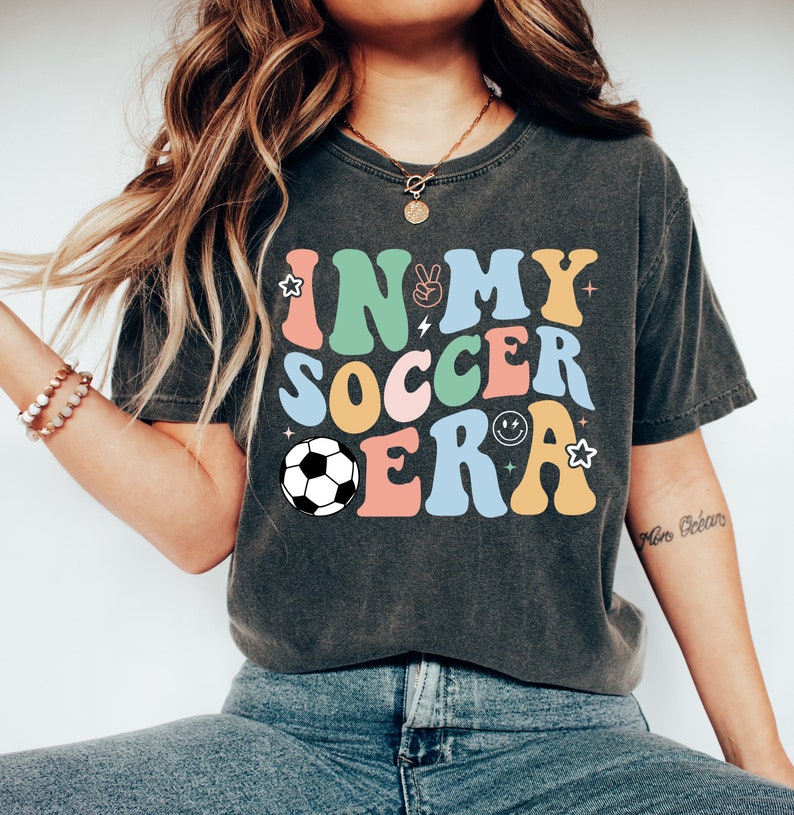 Może przedstawiać: Szary t-shirt z napisem "IN MY SOCCER ERA" w kolorowych, retro literach. W projekcie znajduje się piłka nożna i inne elementy graficzne.