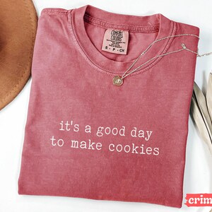 May include: A red t-shirt with the text "it's a good day to make cookies" printed in white. The shirt is made of a soft, comfortable material and has a relaxed fit.