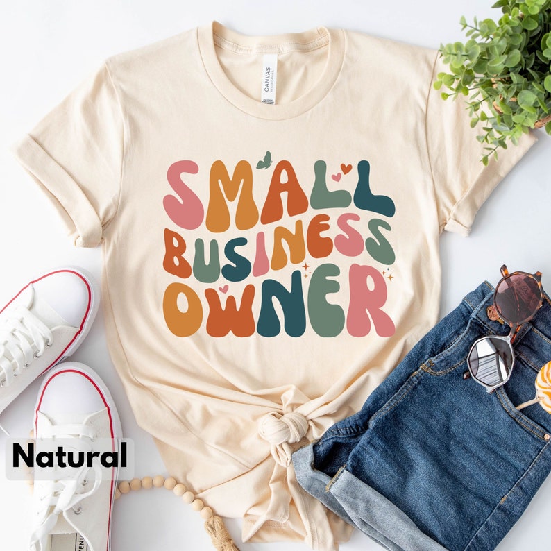 May include: A beige t-shirt with the text "SMALL BUSINESS OWNER" in colorful, retro-style lettering. The shirt is laid flat on a white surface with a pair of white sneakers and a pair of blue denim shorts.