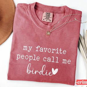 May include: Crimson t-shirt with the text "my favorite people call me birdie" in white script. A gold necklace with a pendant rests on the shirt. A brown hat and dried leaves are in the background.