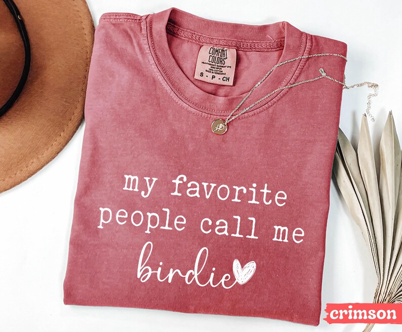 May include: Crimson t-shirt with the text "my favorite people call me birdie" in white script. A gold necklace with a pendant rests on the shirt. A brown hat and dried leaves are in the background.
