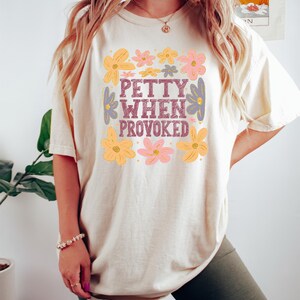 May include: Cream-colored t-shirt with the phrase "PETTY WHEN PROVOKED" in a floral design. The shirt features a square of flowers in yellow, pink, and purple. The shirt is a casual, comfortable style.