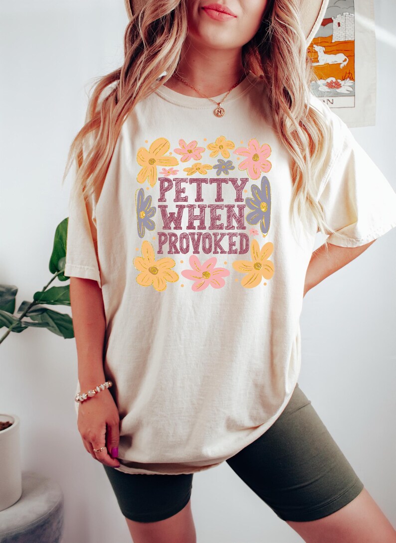 May include: Cream-colored t-shirt with the phrase "PETTY WHEN PROVOKED" in a floral design. The shirt features a square of flowers in yellow, pink, and purple. The shirt is a casual, comfortable style.