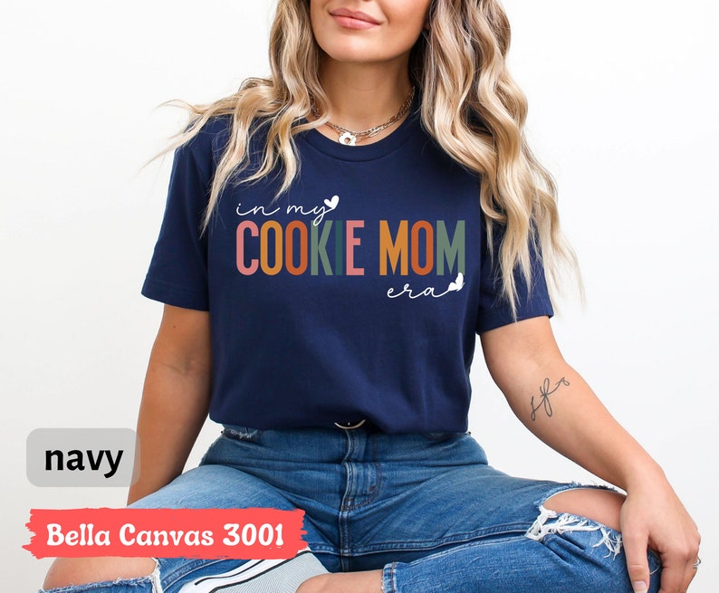 May include: A navy blue t-shirt with the text "in my cookie mom era" in colorful letters. The shirt is made by Bella Canvas and is a 3001 style.