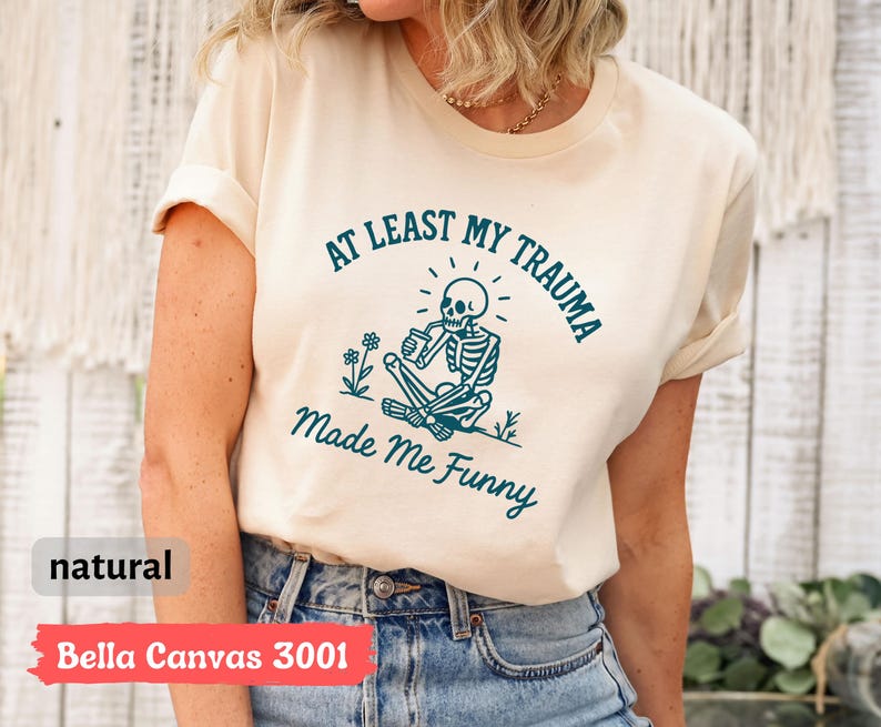K&ouml;nnte beinhalten: Ein naturfarbenes Bella Canvas 3001 T-Shirt mit dem Text "AT LEAST MY TRAUMA Made Me Funny" in Teal. Die Grafik zeigt ein Skelett, das aus einer Tasse trinkt. Das Shirt ist ein Freizeitkleidungsst&uuml;ck.