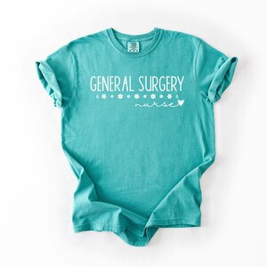 Może przedstawiać: Turkusowa koszulka z napisem "GENERAL SURGERY nurse" w białym kolorze. Tekst jest ozdobiony małymi białymi grafikami kwiat&oacute;w.