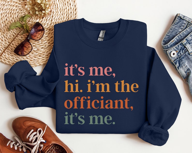 May include: Navy blue sweatshirt with the text "it's me, hi. i'm the officiant, it's me." in colourful lettering.